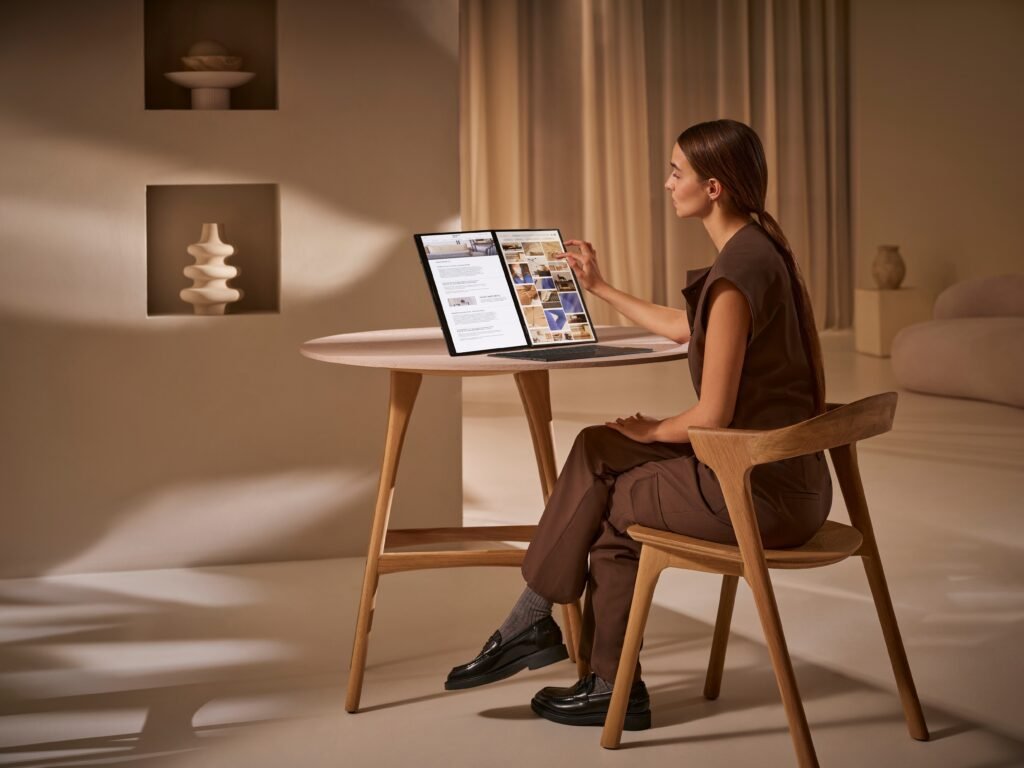A person seated at a round wooden table touches the second screen of a Zenbook DUO standing in desktop mode